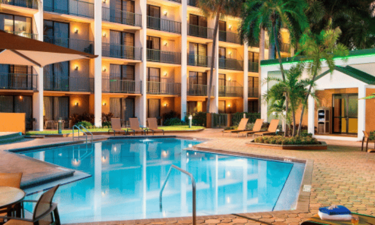 Hotel courtyard at night with lit pool, lounge chairs, palms, and building balconies.