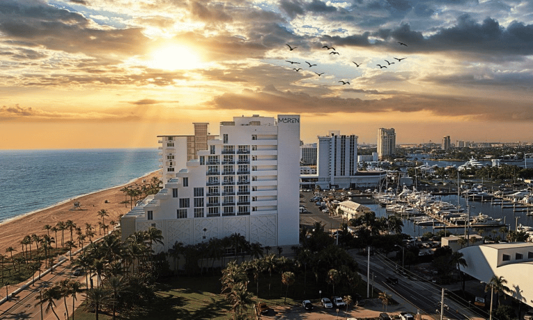 Coastal hotel at sunrise, with palm trees, ocean, birds, and cityscape in the background.