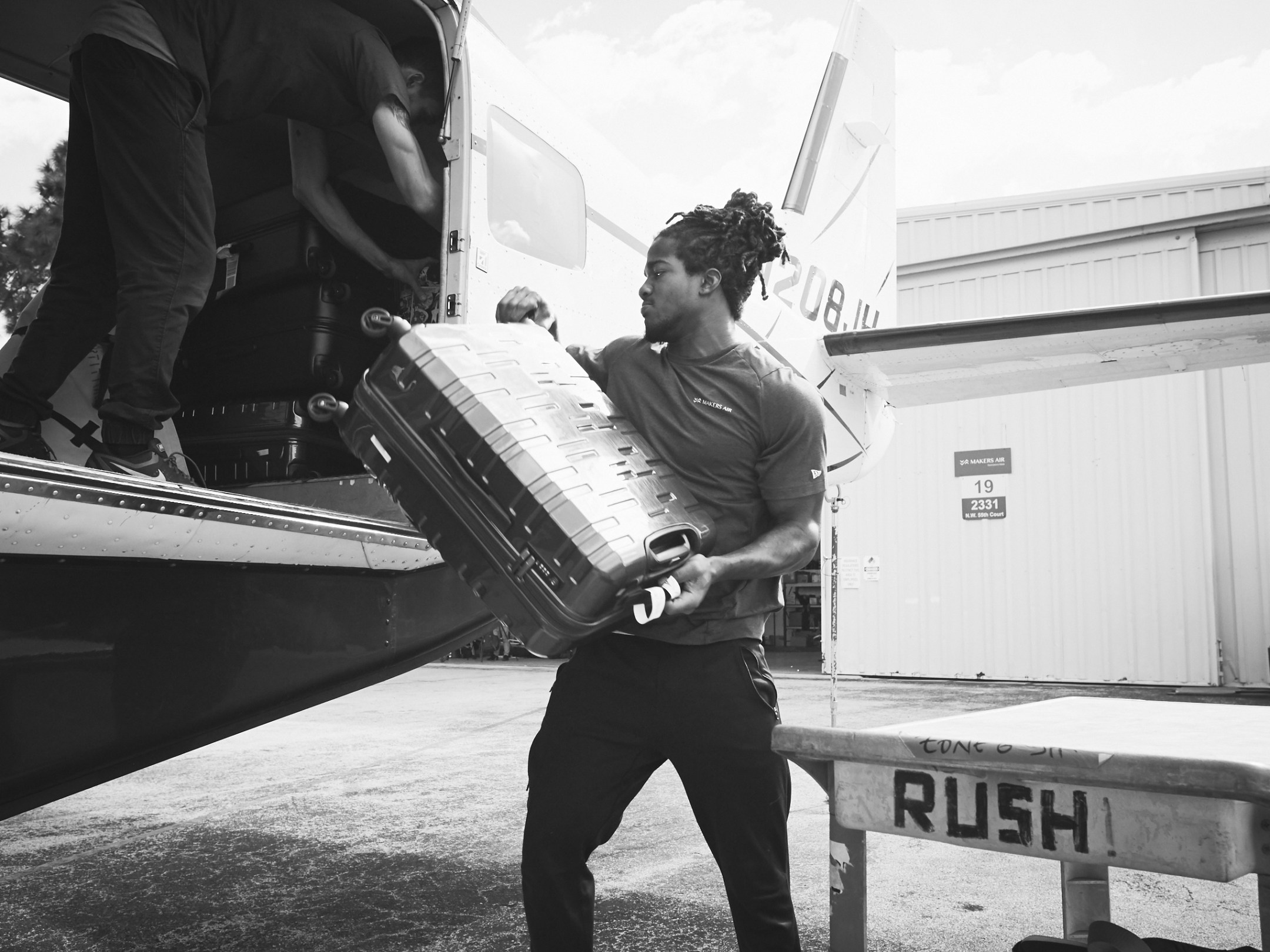 Two people loading a suitcase onto a small aircraft outside a hangar.