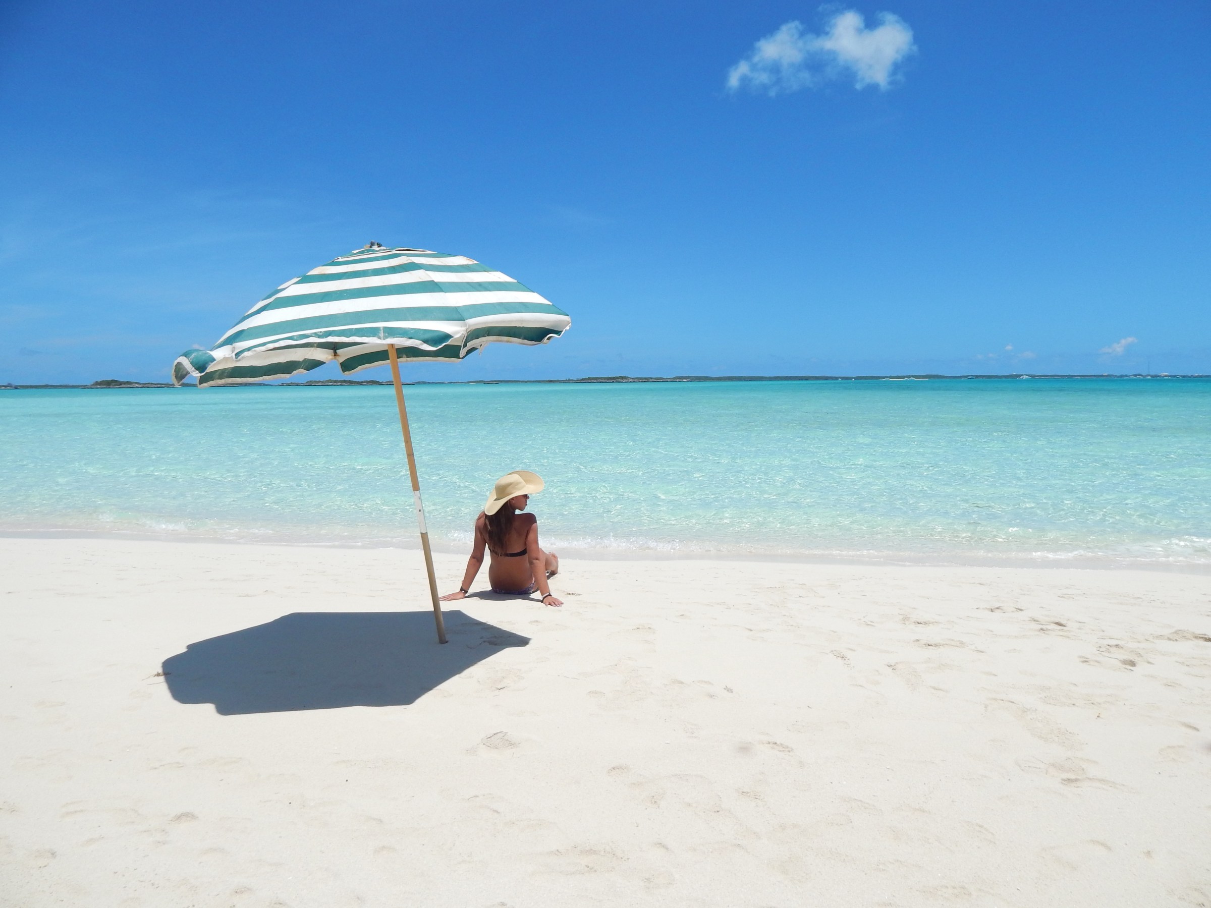 Person under blue-striped umbrella on a sandy beach facing clear turquoise water.