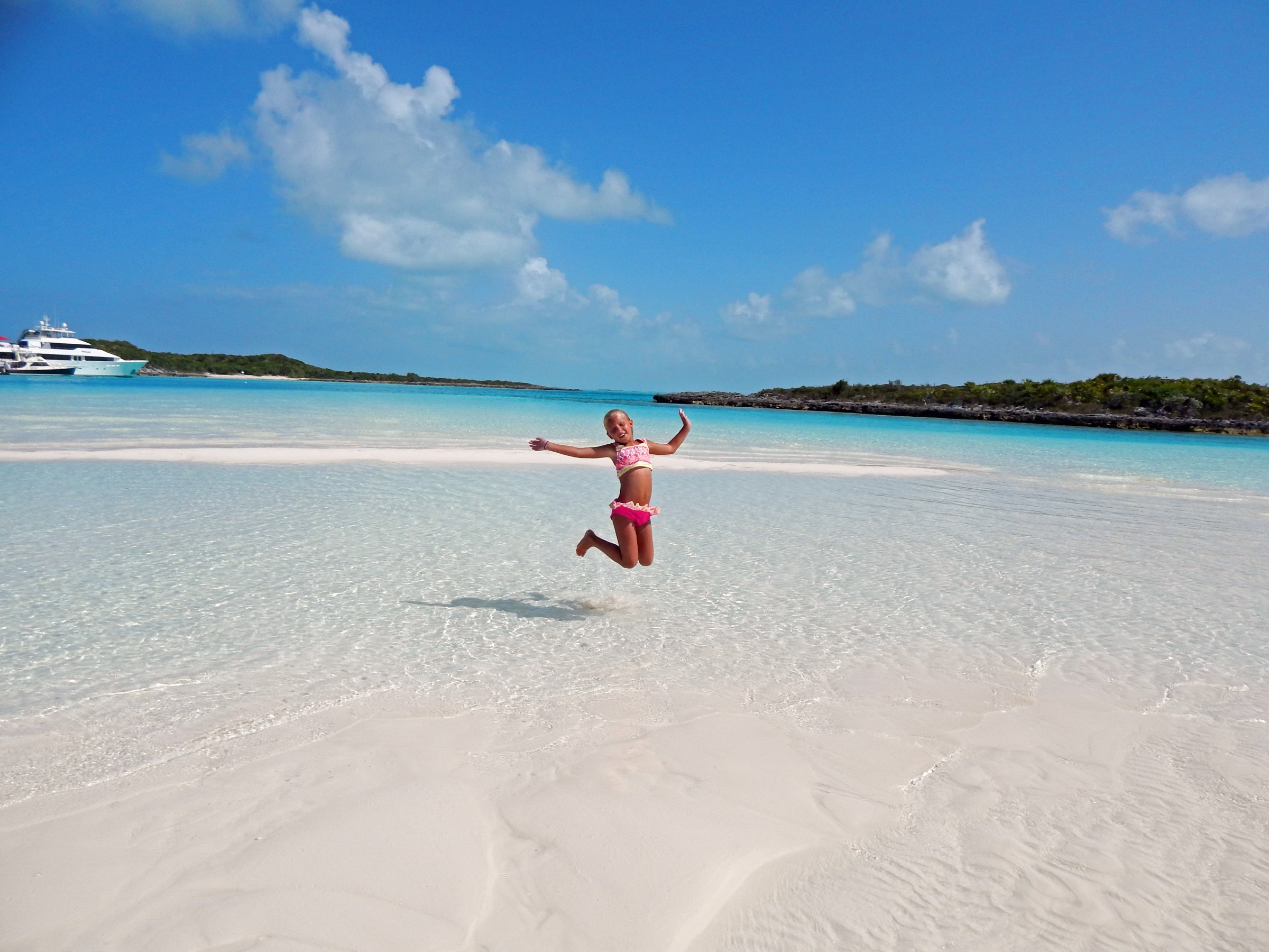 Child in pink swimsuit jumps on tropical beach with clear water and yachts in background.