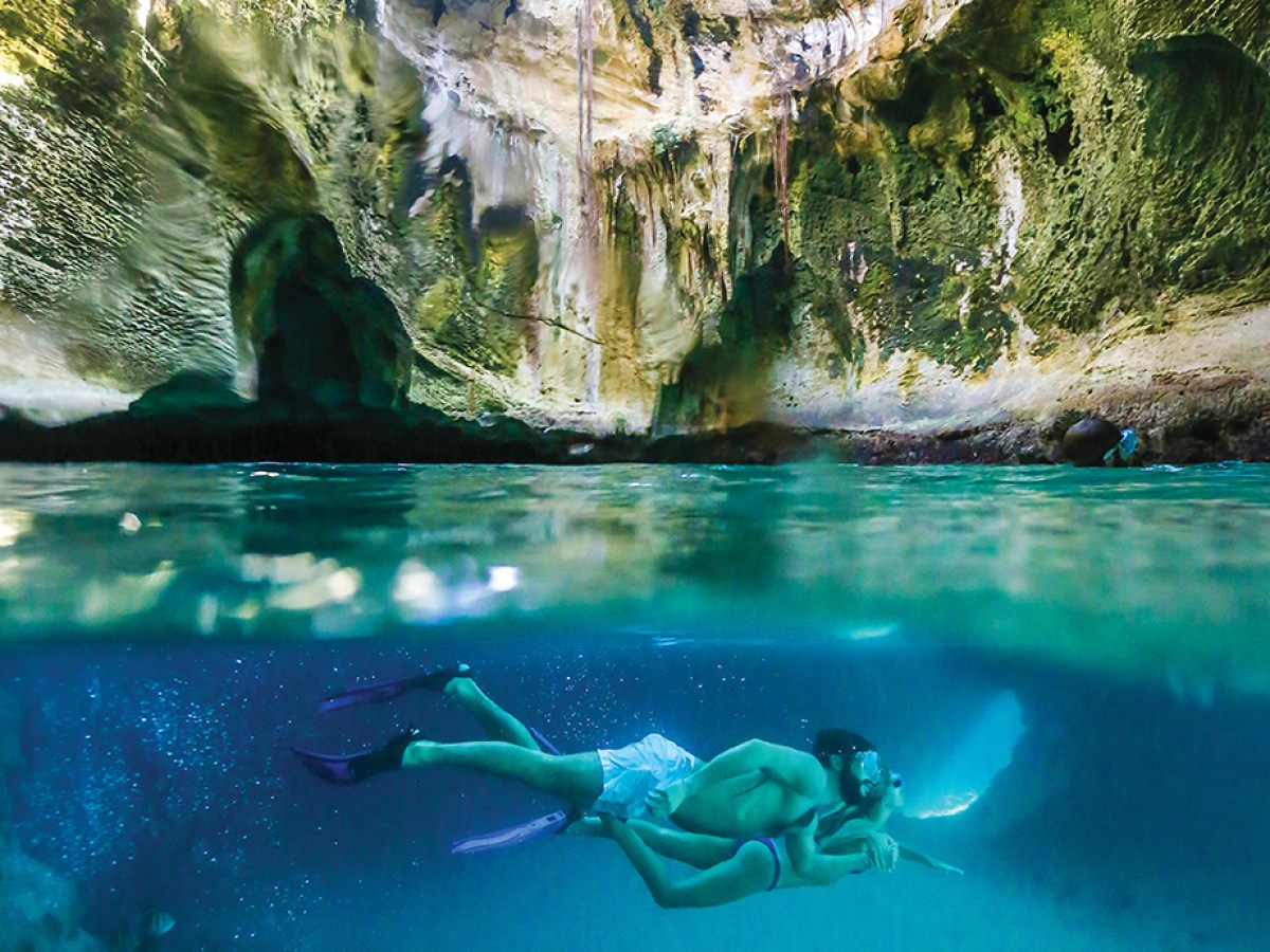 Man snorkeling underwater in a cave with clear blue water and sunlit ceiling.