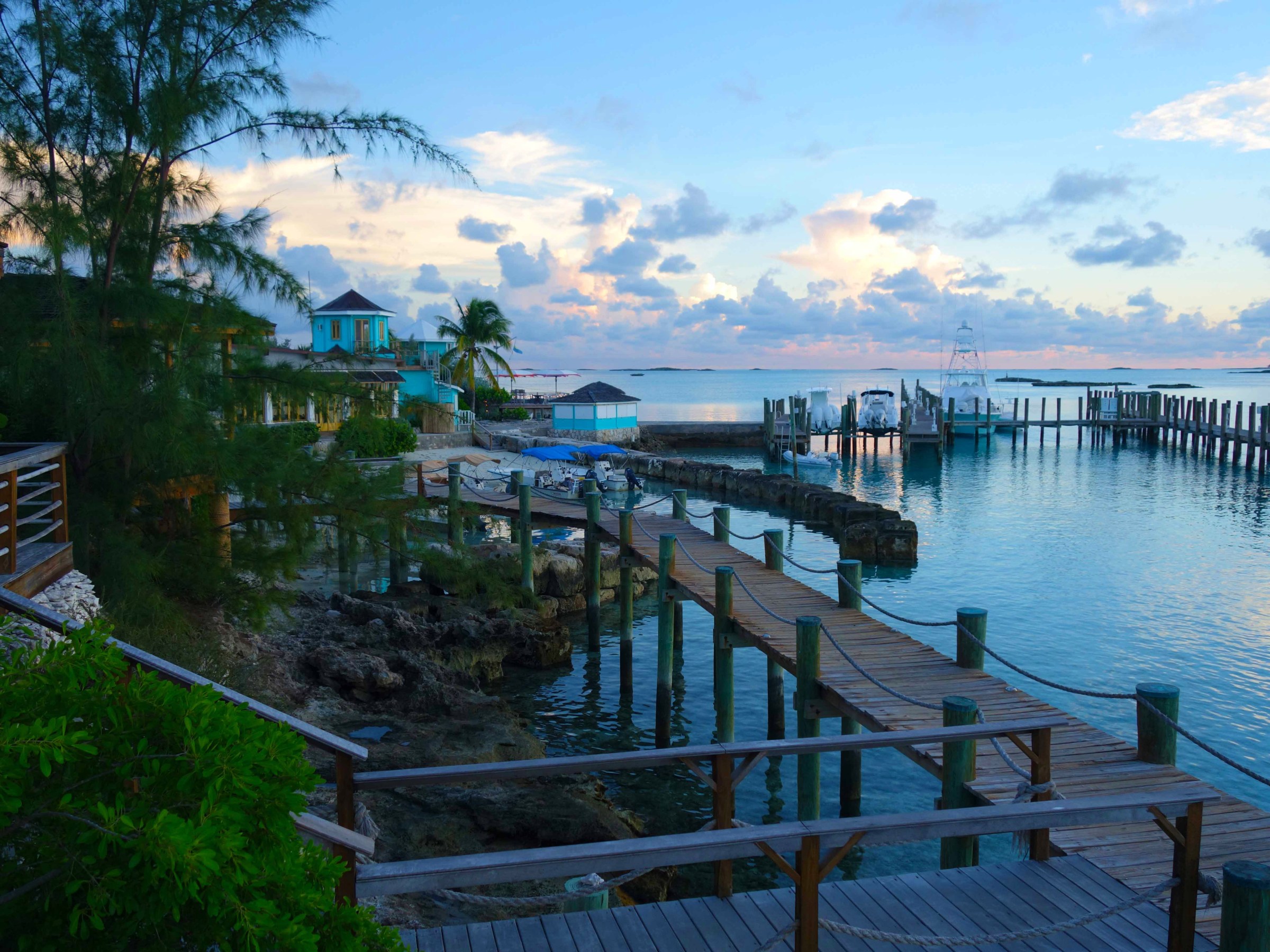 Coastal scene with wooden docks, turquoise buildings, palm trees, and boats at sunset.
