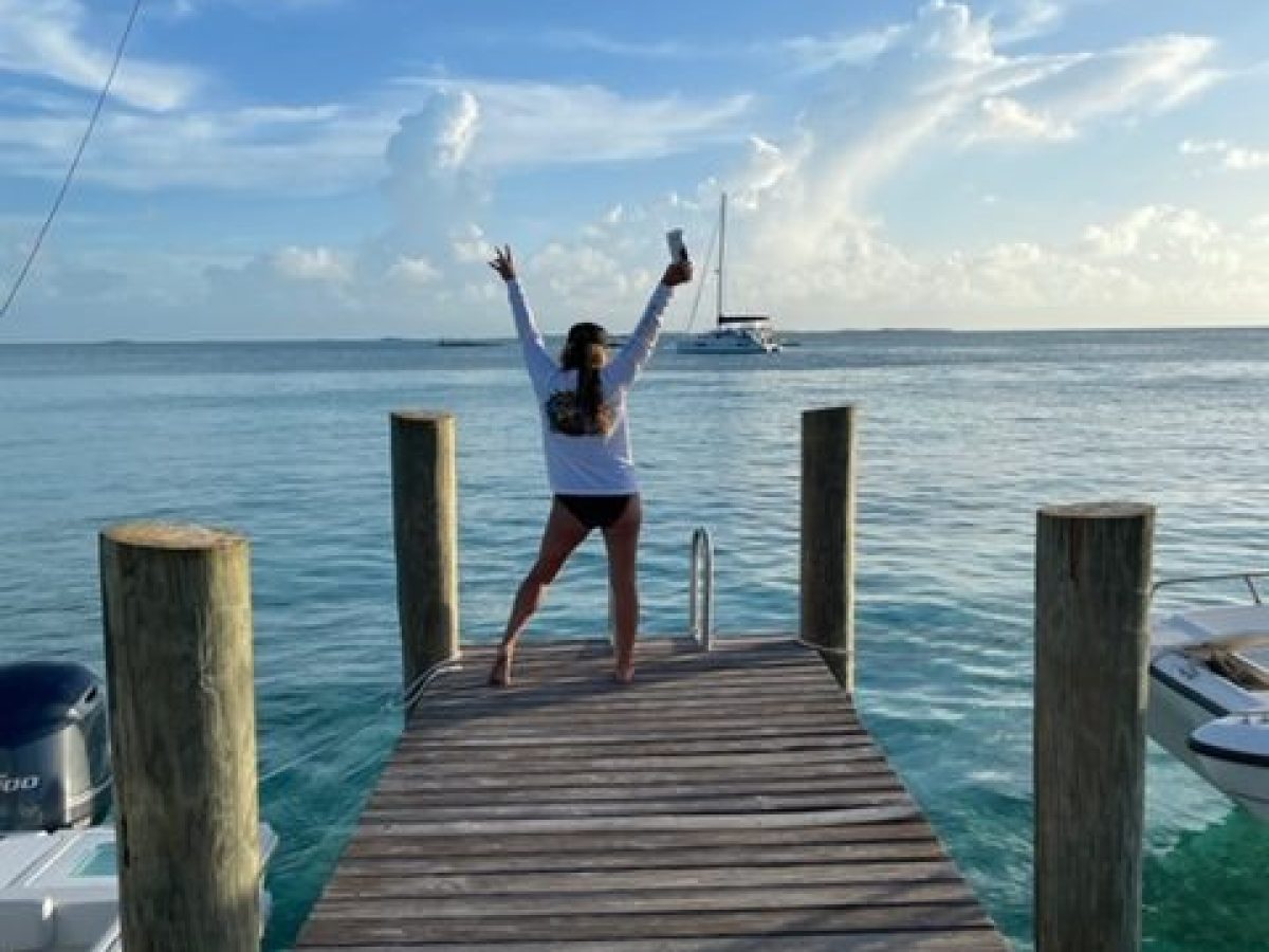 Person on dock, arms raised, facing ocean with a boat in the distance under a partly cloudy sky.