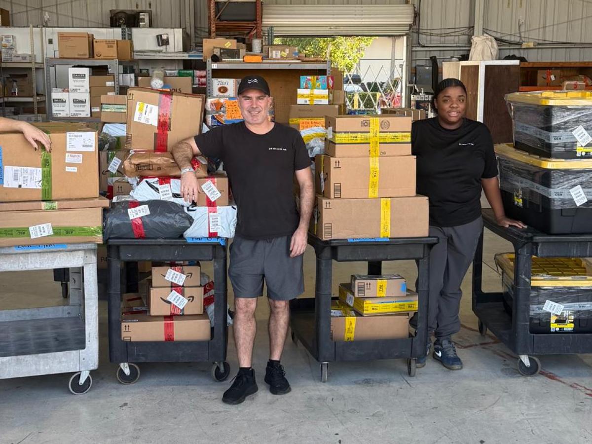 Four people in a warehouse with carts full of packages, smiling at the camera.