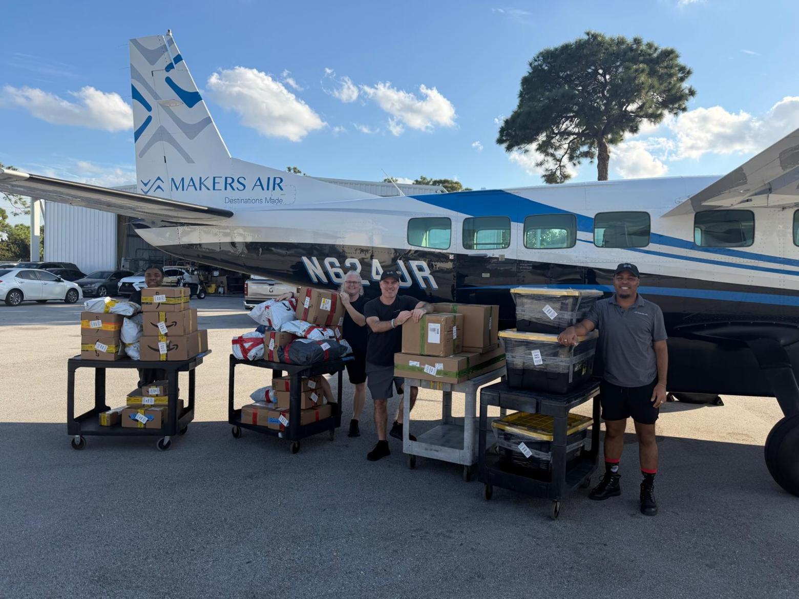 Three people stand by a small airplane with carts full of packages on a sunny day.