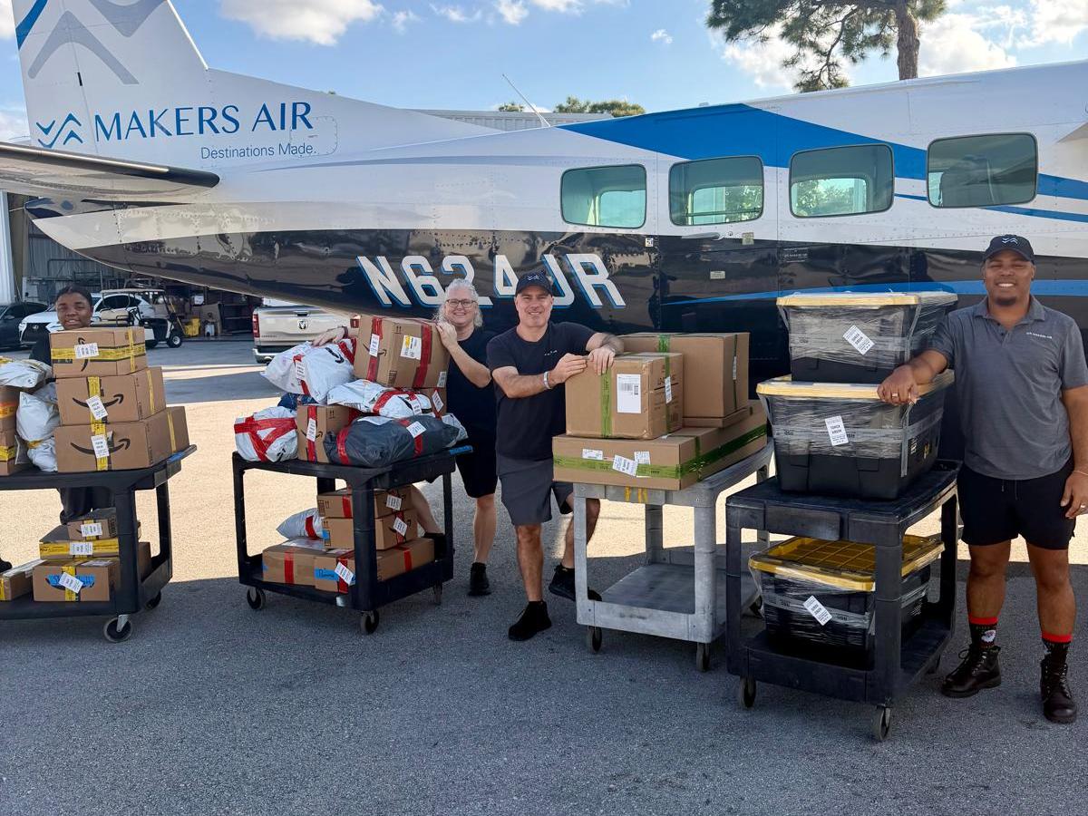 Three people standing by an airplane with carts of packages under a blue sky.