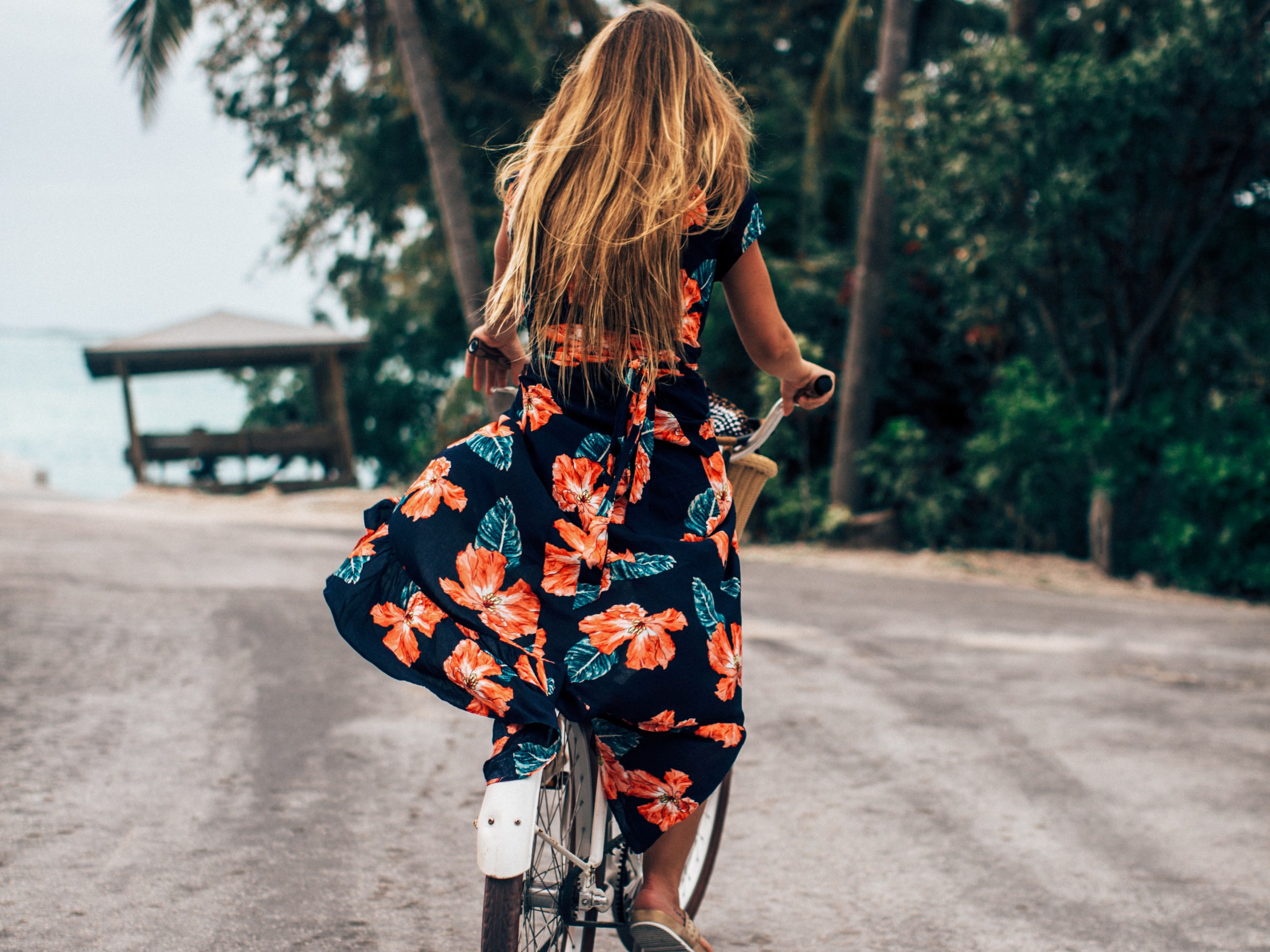 Woman with long hair rides bike in floral dress on road with palm trees.