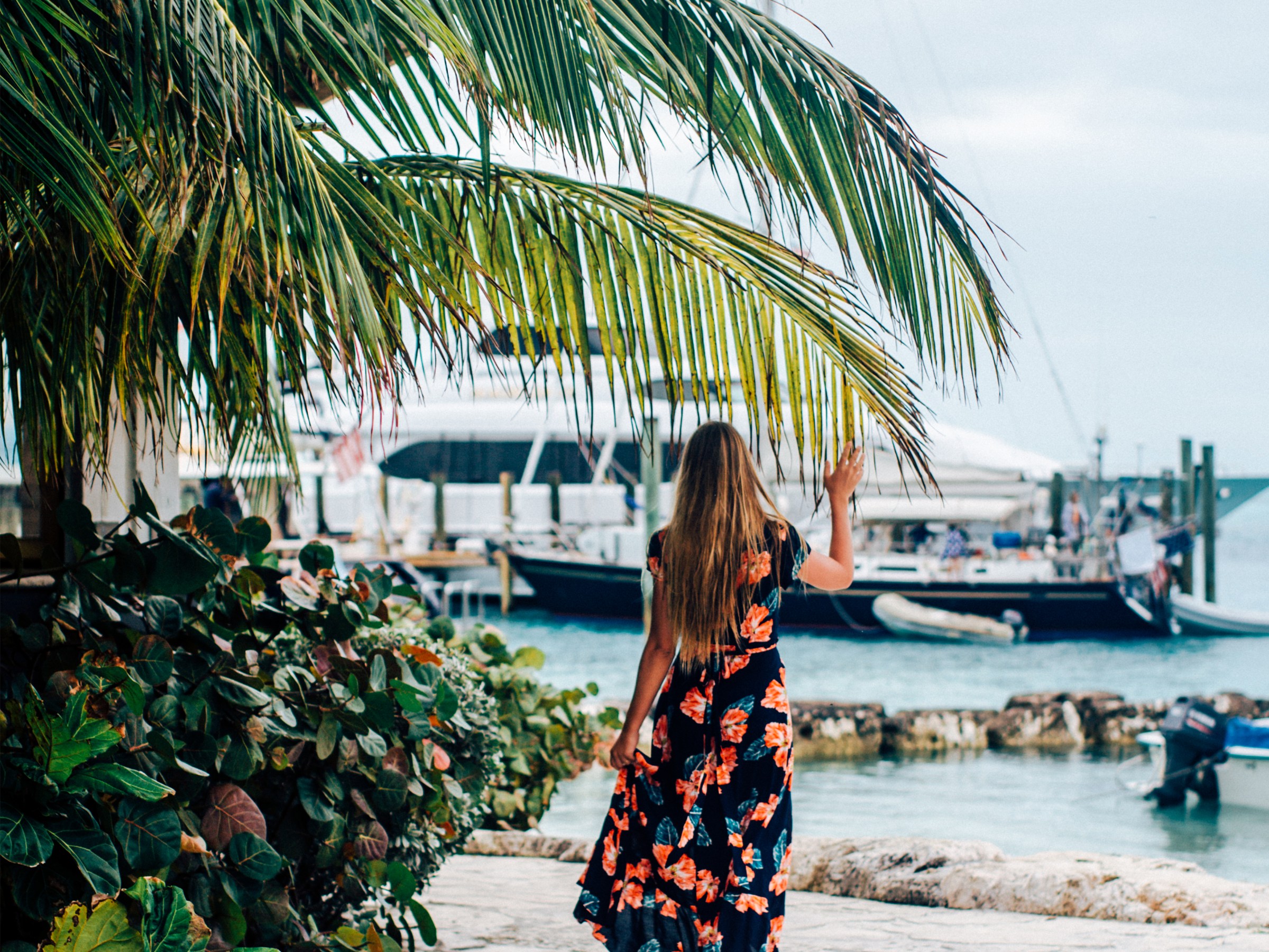 Woman in floral dress walks along palm-lined path by a marina.