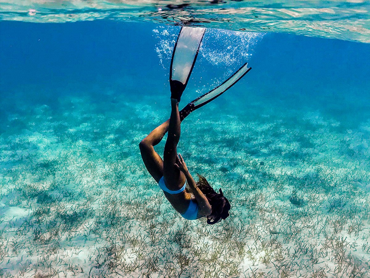 Person snorkeling underwater with fins in clear blue sea and sandy seabed.