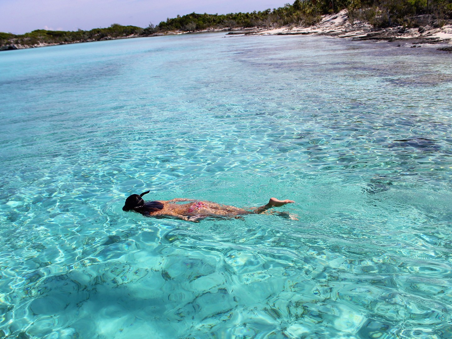 Person snorkeling in clear turquoise water near a tropical shoreline.