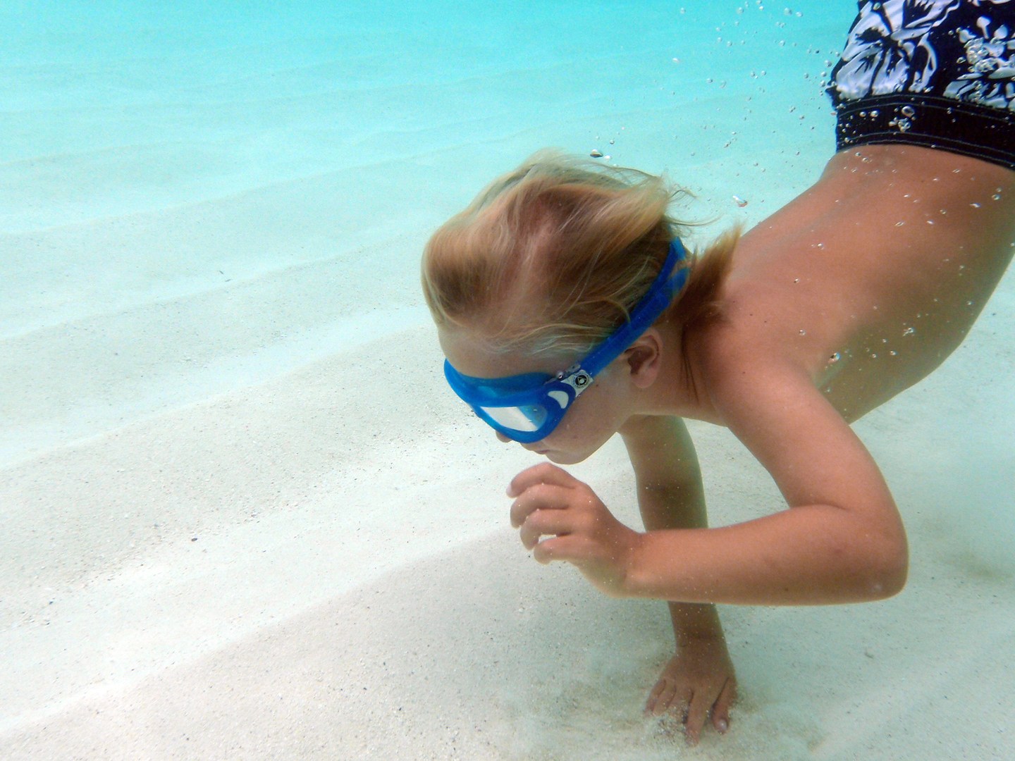 Child underwater wearing blue goggles, swimming on sandy pool floor.