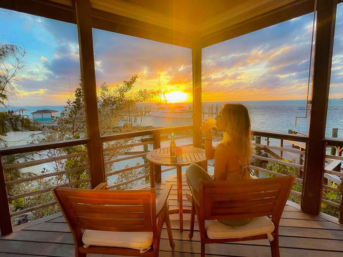 Person on deck drinking, watching ocean sunset by wooden table and chairs.