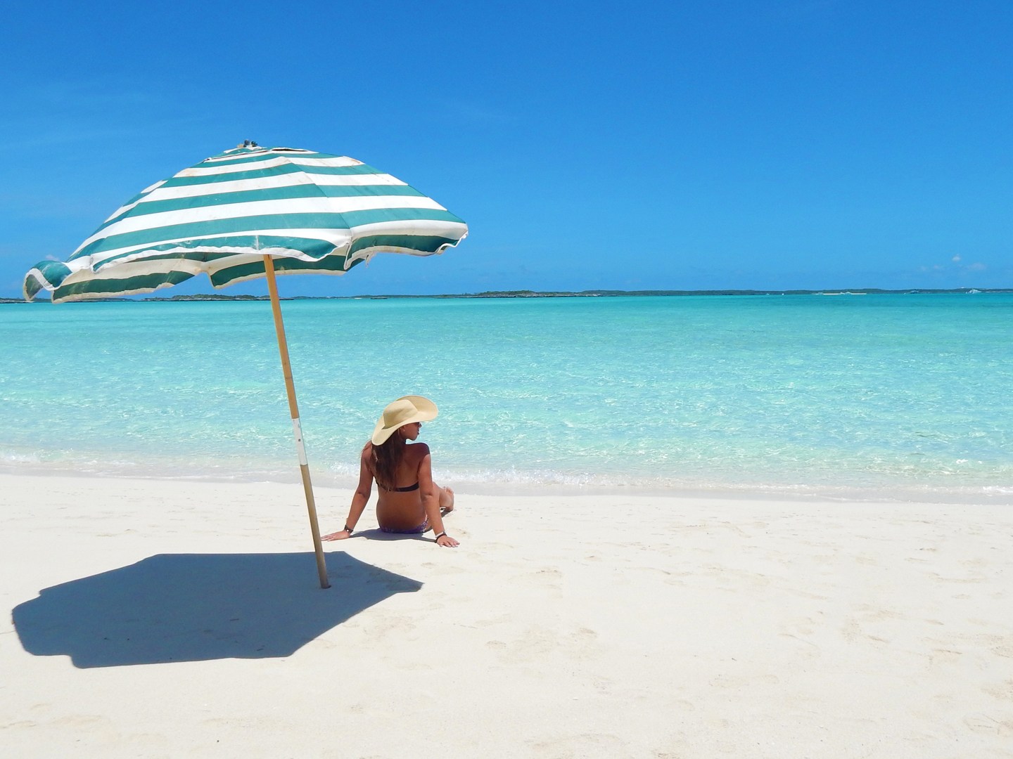 Person sitting on a beach under a striped umbrella, facing clear turquoise water.