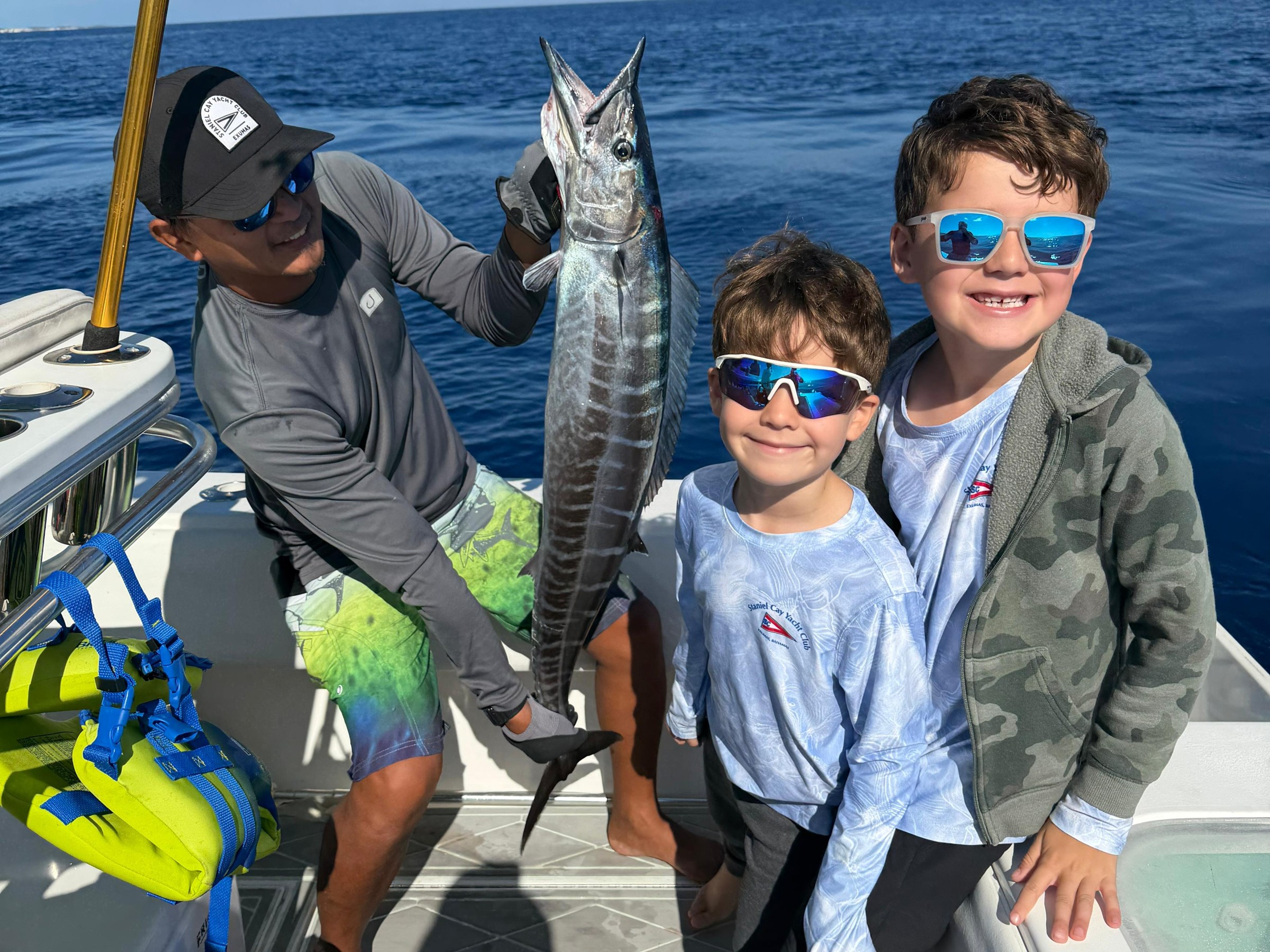 Three people on a boat; one holds a large fish, two kids wear sunglasses and smile.