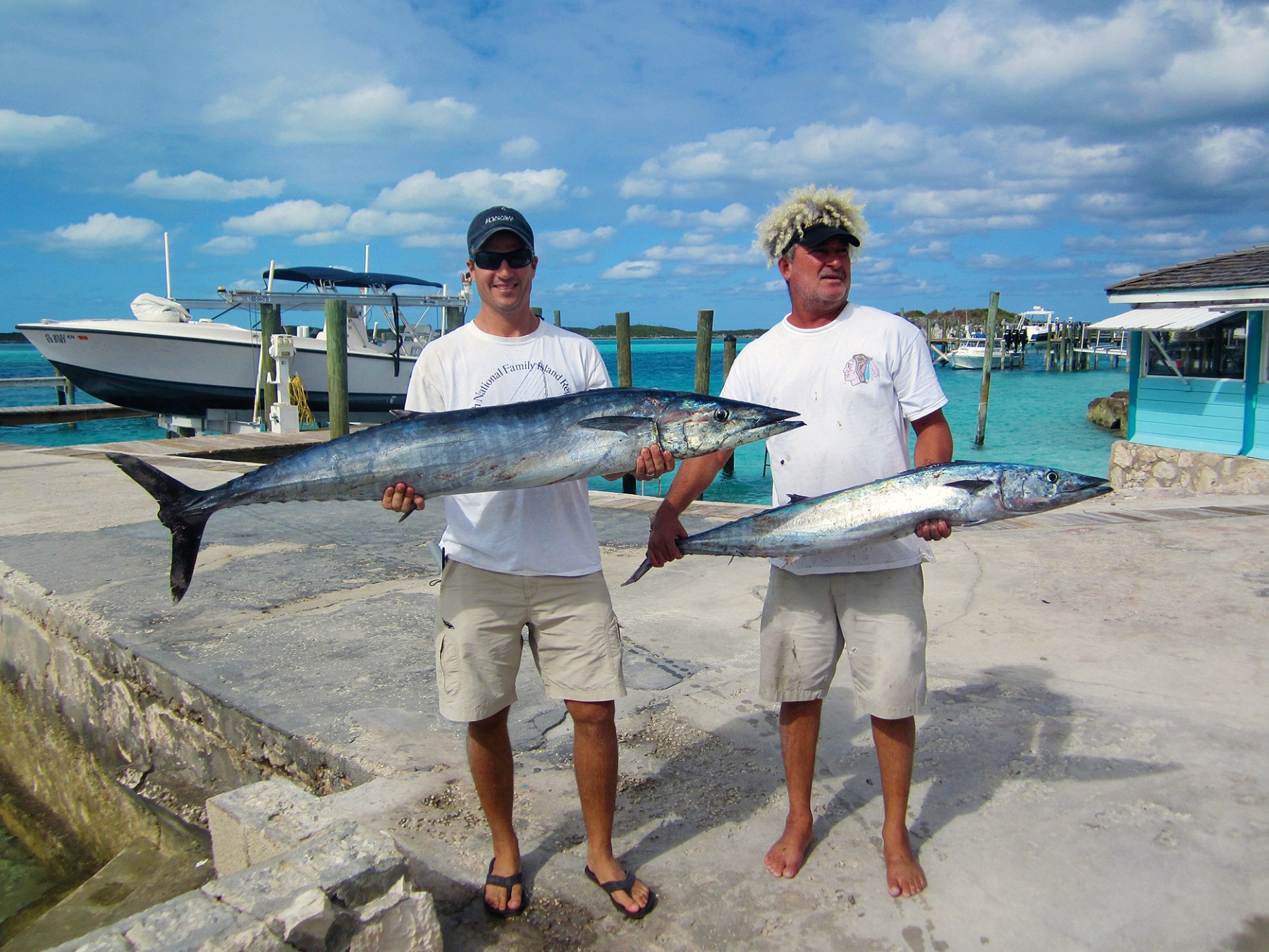 Two men holding large fish on a dock by the sea, with boats in the background.
