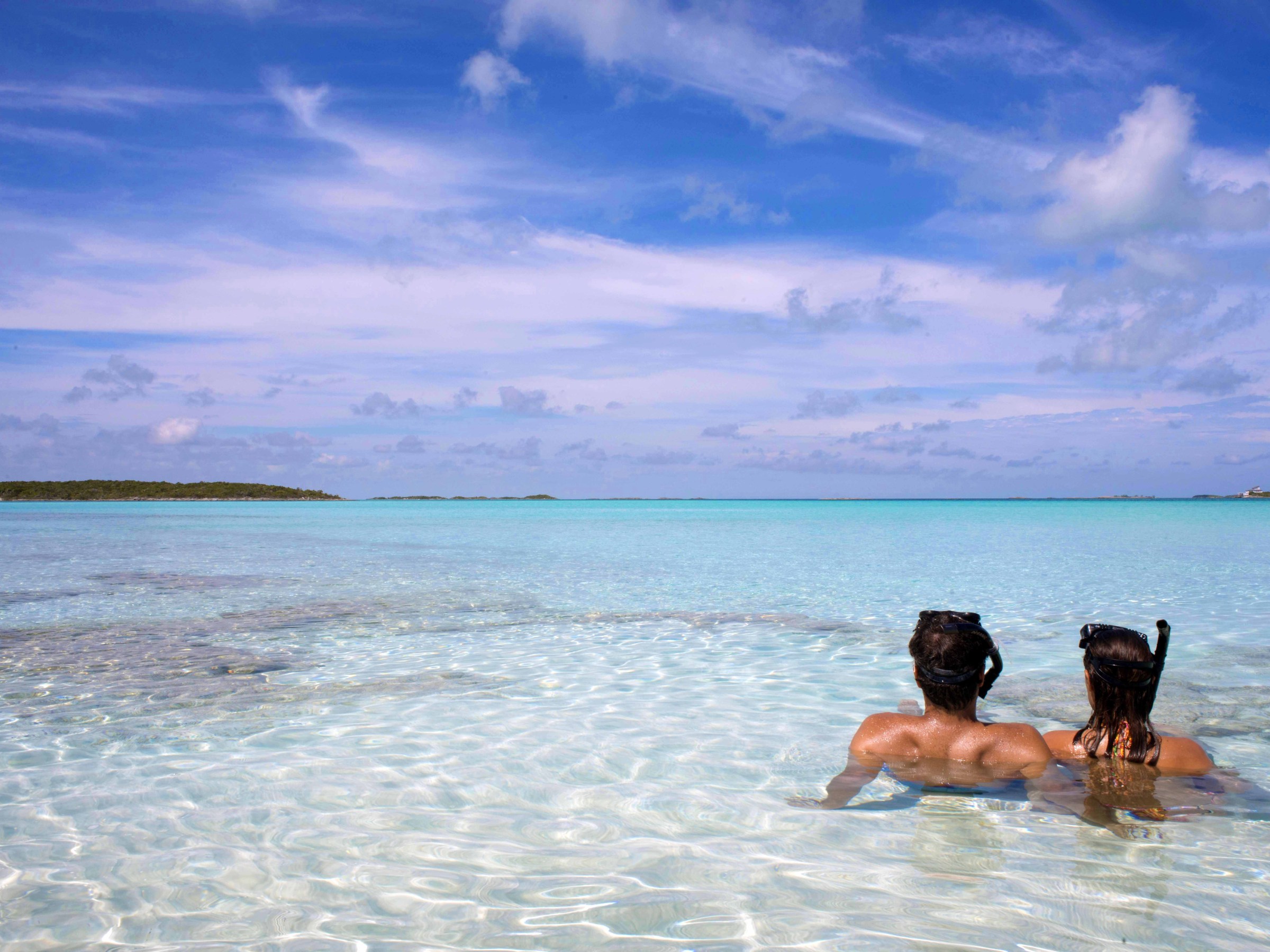 Two people in clear shallow water with snorkels facing the horizon under a blue sky.