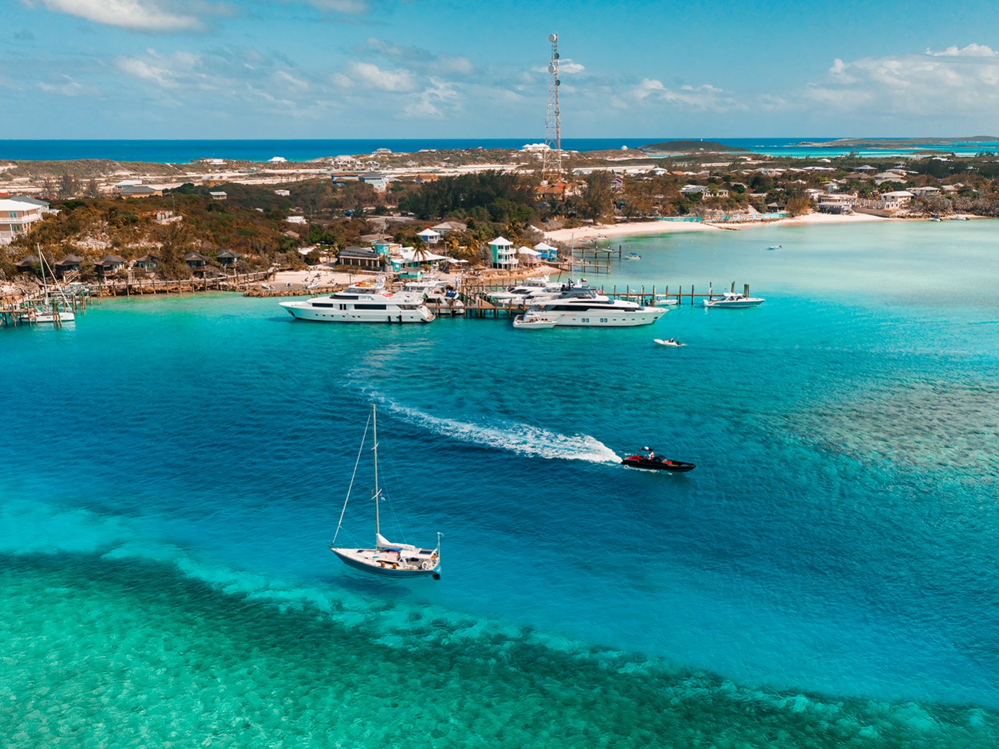Aerial view of boats in a turquoise bay with a small marina and buildings on the shoreline.